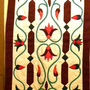 May include: A white fabric wall hanging with a repeating pattern of stylized flowers in shades of red, green, and brown. The flowers are outlined in green and have a diamond pattern in the center. The fabric is bordered with a dark brown fabric.