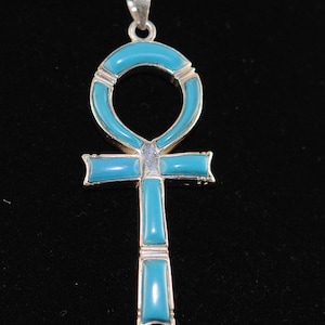 May include: A silver-toned ankh pendant with turquoise-colored inlays. The ankh, a symbol of life, features a loop at the top, a horizontal bar, and a vertical bar. The pendant is suspended from a delicate silver chain.