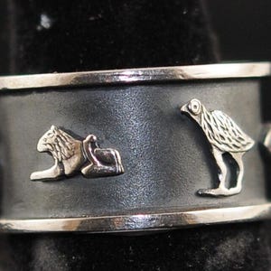 May include: Silver ring with a dark gray band featuring raised silver relief designs. The designs include a lion and a bird. The ring has a polished silver edge.