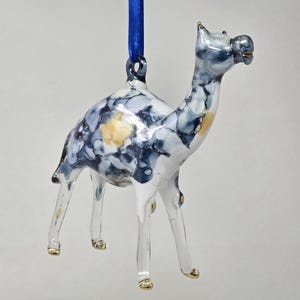 May include: A glass camel ornament featuring a marbled pattern of white, dark blue, and light yellow swirls. The camel has clear glass legs with gold-painted hooves and a blue satin ribbon loop attached to the top for hanging purposes.