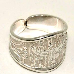 May include: A silver-colored ring with an engraved Egyptian design. The ring features hieroglyphs and a winged figure. The band is wide and has a smooth, polished finish. The ring is a statement piece.