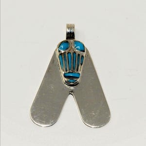 May include: Silver-tone pendant with a unique design. The pendant features a central section with turquoise-colored inlays and a stylized shape. The pendant has a loop at the top for a chain.