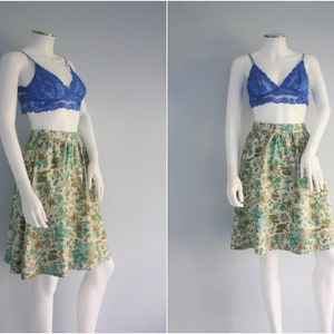 May include: A white skirt with a floral print in shades of green, brown, and blue. The skirt has a fitted waist and a flared hem. It is paired with a blue lace bralette.