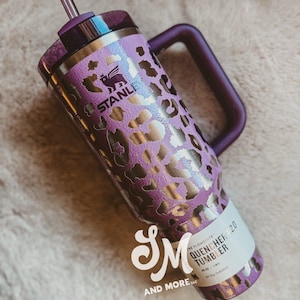 May include: A purple and gold leopard print Stanley Quencher 2.0 tumbler with a handle and straw. The tumbler has a purple lid and handle. The text "STANLEY" is visible. Designed for all-day hydration, it is a stylish drinks container.