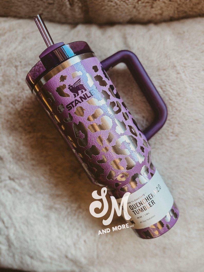 May include: A purple and gold leopard print Stanley Quencher 2.0 tumbler with a handle and straw. The tumbler has a purple lid and handle. The text "STANLEY" is visible. Designed for all-day hydration, it is a stylish drinks container.