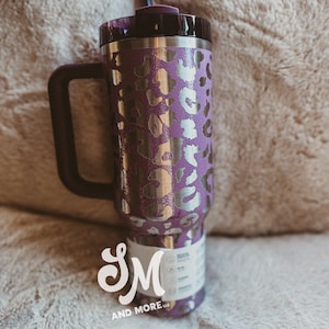 May include: Purple travel mug with a silver leopard print pattern, a black handle, and a black lid. The mug has a straw and a white label with the text "SM AND MORE LLC".