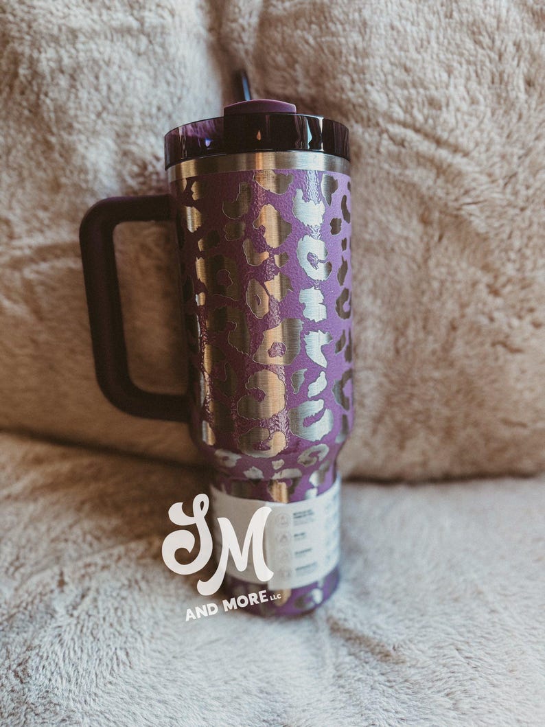 May include: Purple travel mug with a silver leopard print pattern, a black handle, and a black lid. The mug has a straw and a white label with the text "SM AND MORE LLC".