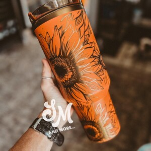 May include: Orange tumbler with a black sunflower design. The tumbler has a gold-colored rim and a straw. The design features detailed black outlines of sunflowers on the orange background. The tumbler is held in a hand.