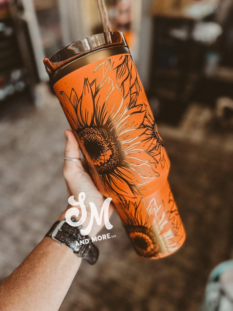 May include: Orange tumbler with a black sunflower design. The tumbler has a gold-colored rim and a straw. The design features detailed black outlines of sunflowers on the orange background. The tumbler is held in a hand.