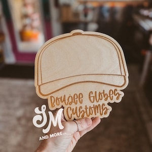 May include: A wooden sign in the shape of a baseball cap. The sign has the words "Bousee Globes Customs" and "SM AND MORE LLC" engraved on it. The sign is held by a person.