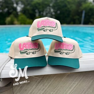 May include: Three beige baseball caps with teal green brims. Each cap features the word "LATER" in pink above a cartoon alligator. The caps are displayed near a pool. The caps are from "SM AND MORE LLC".