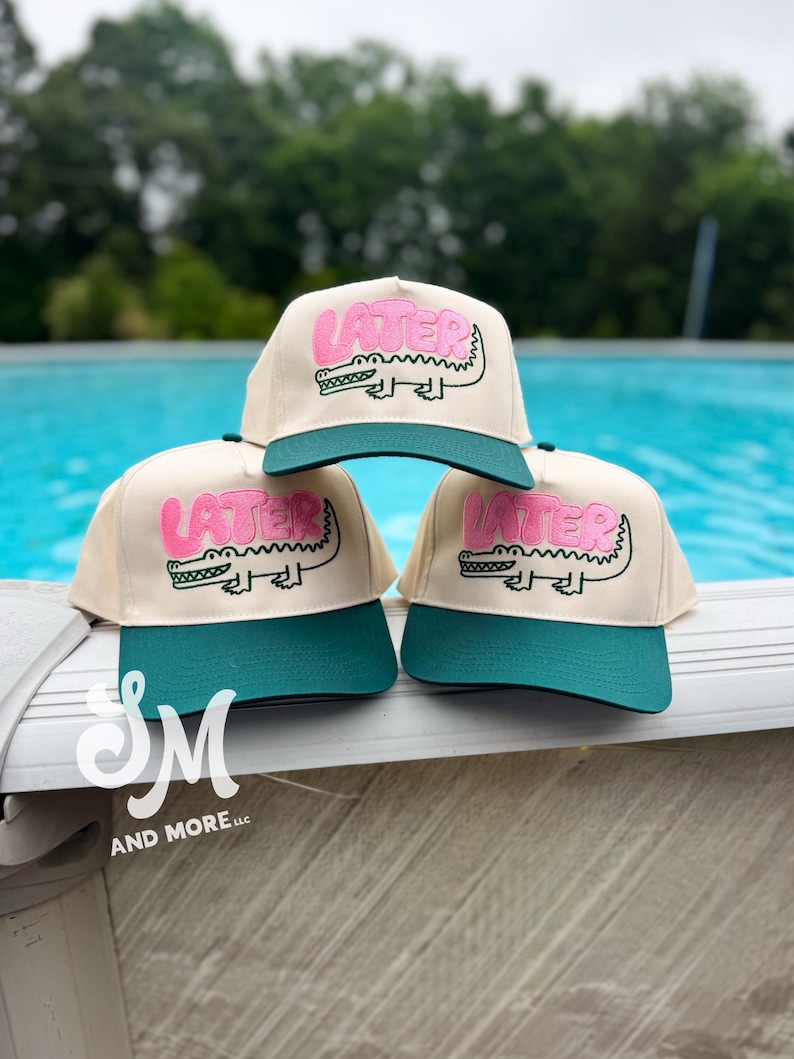 May include: Three beige baseball caps with teal green brims. Each cap features the word "LATER" in pink above a cartoon alligator. The caps are displayed near a pool. The caps are from "SM AND MORE LLC".