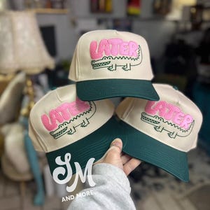 May include: Three cream-colored baseball caps with dark green brims. Each cap features the word "LATER" in pink above a pink alligator illustration. The caps are a fun, casual accessory.