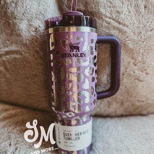 May include: Purple and silver Stanley Quencher H2.0 Tumbler with a handle and straw. The tumbler features a leopard print design and the Stanley logo. The bottom of the tumbler has a white label with the text "QUENCHER H2.0 TUMBLER".