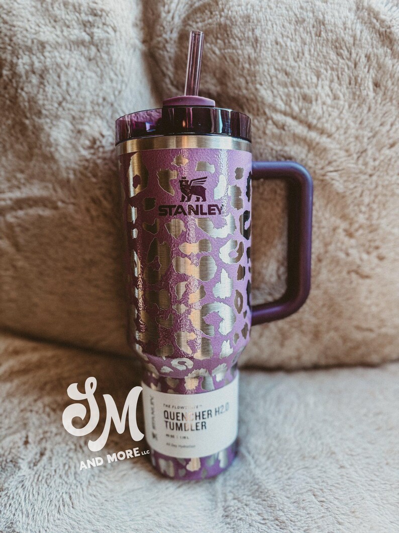 May include: Purple and silver Stanley Quencher H2.0 Tumbler with a handle and straw. The tumbler features a leopard print design and the Stanley logo. The bottom of the tumbler has a white label with the text "QUENCHER H2.0 TUMBLER".