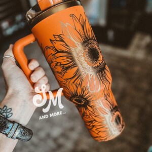 May include: Orange tumbler with a black and gold sunflower design. The tumbler has a handle and a clear lid with an orange top. The text "SM AND MORE LLC" is visible on the tumbler.