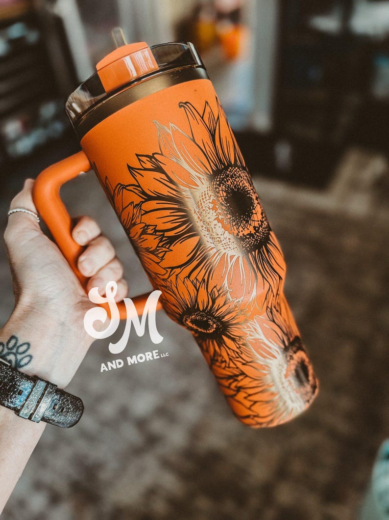 May include: Orange tumbler with a black and gold sunflower design. The tumbler has a handle and a clear lid with an orange top. The text "SM AND MORE LLC" is visible on the tumbler.