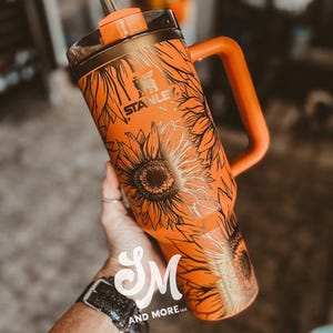 May include: Orange Stanley tumbler with a black sunflower design. The tumbler has a handle, a straw, and a black lid. The Stanley logo is visible on the front. The tumbler is held by a person.
