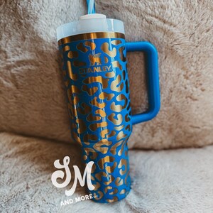 May include: A blue Stanley tumbler with a gold leopard print pattern. The tumbler features a blue handle, a white lid, and a blue and white striped straw. The Stanley logo is visible on the front.