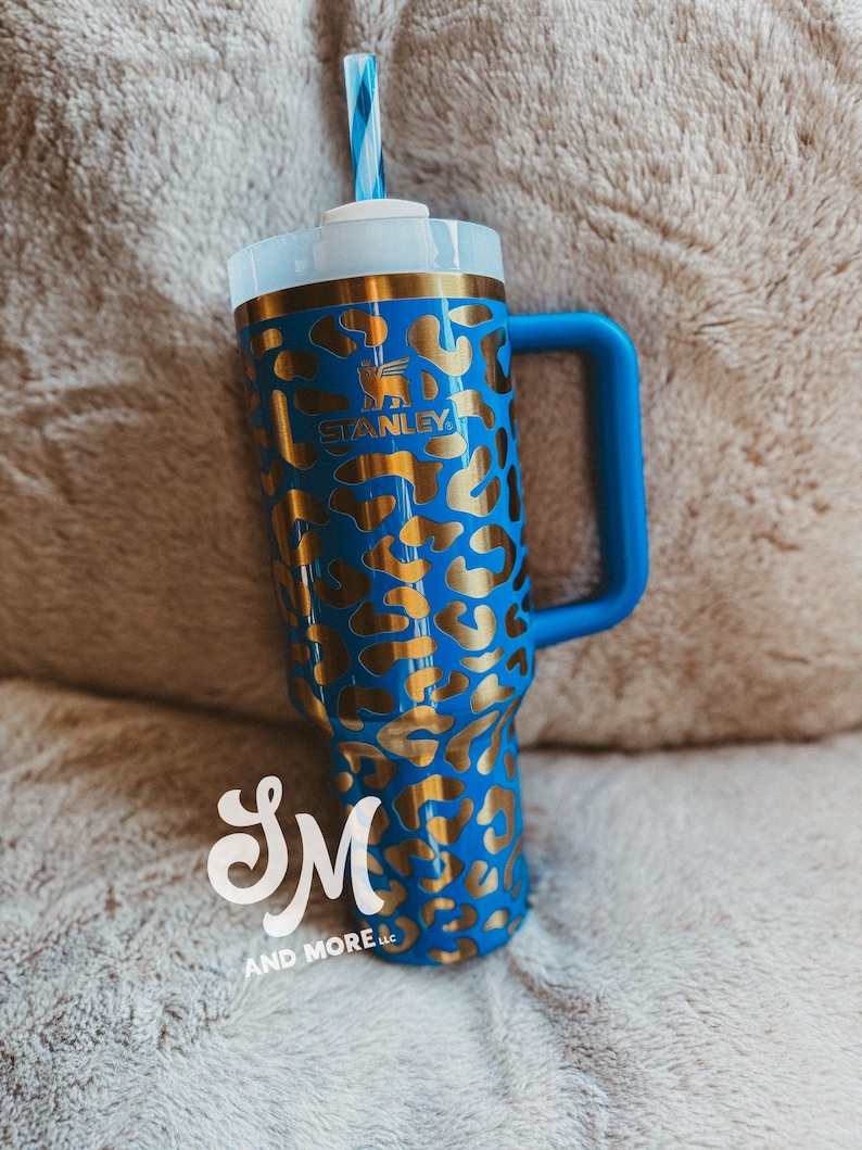 May include: A blue Stanley tumbler with a gold leopard print pattern. The tumbler features a blue handle, a white lid, and a blue and white striped straw. The Stanley logo is visible on the front.