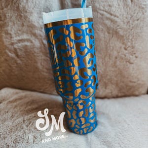May include: A blue tumbler with a gold leopard print pattern. The tumbler features a white lid, a gold rim, and a blue and white striped straw. The logo "SM AND MORE llc" is visible at the bottom.