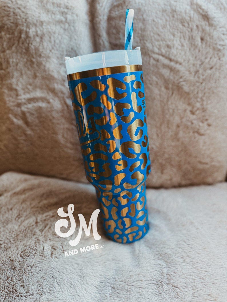 May include: A blue tumbler with a gold leopard print pattern. The tumbler features a white lid, a gold rim, and a blue and white striped straw. The logo "SM AND MORE llc" is visible at the bottom.
