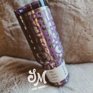 May include: A purple stainless steel travel mug with a silver leopard print pattern. The mug has a matching purple lid and a clear straw. The logo "SM AND MORE llc" is visible on the bottom of the mug.
