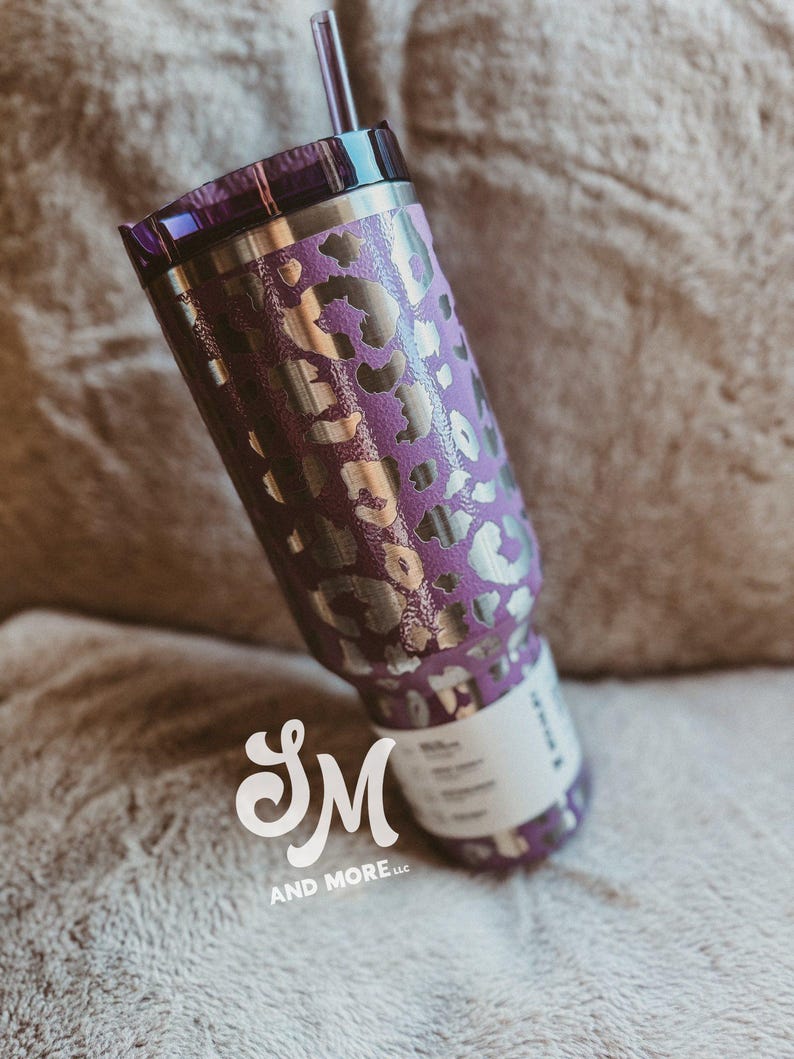 May include: A purple stainless steel travel mug with a silver leopard print pattern. The mug has a matching purple lid and a clear straw. The logo "SM AND MORE llc" is visible on the bottom of the mug.