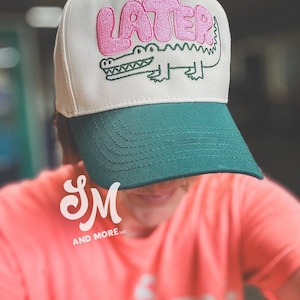 May include: A baseball cap with a cream-colored crown and a green bill. The cap features the word "LATER" in pink bubble letters above a green alligator illustration. The cap is a stylish accessory.