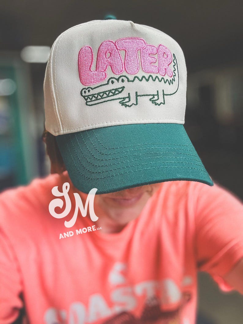May include: A baseball cap with a cream-colored crown and a green bill. The cap features the word "LATER" in pink bubble letters above a green alligator illustration. The cap is a stylish accessory.