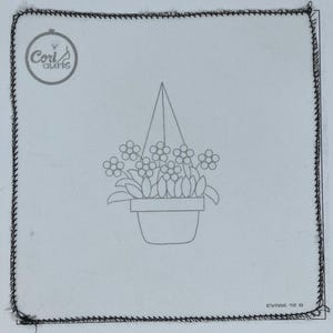 May include: A white square fabric with a black stitched border. The center features a line drawing of a hanging flower pot with flowers and leaves. The top left corner has a circular logo with the text "Cori Crafts".