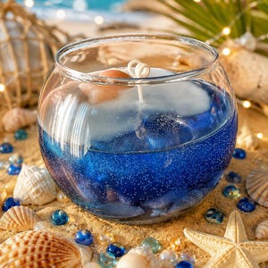 May include: A round glass bowl candle featuring a deep blue gel wax base filled with tiny air bubbles and embedded seashells. The top layer consists of white wax with a centered wick, accented by a small, light-colored seashell resting on the surface.