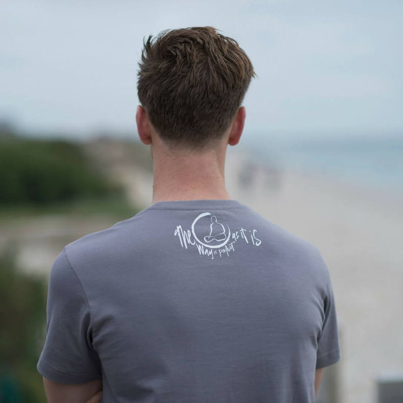 May include: A person wearing a gray t-shirt with a white graphic on the back. The graphic features a circle with a person sitting in a meditative pose and the text "The Way It Is Magic Pocket".