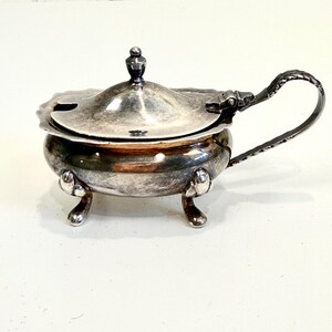 May include: A small, antique silver-plated serving dish with a lid and handle. The dish has a round body, three small feet, and a decorative handle. The lid has a small knob on top. The dish is likely used for serving sauces or condiments.