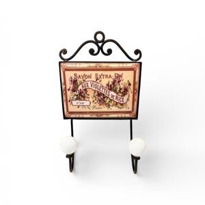 May include: A decorative wall hook with a rectangular sign featuring the text "Savon Extra-Fin Aux Violettes de Nice Paris." The sign has a floral design in shades of purple, pink, and brown. The hook is black with two white ceramic knobs.