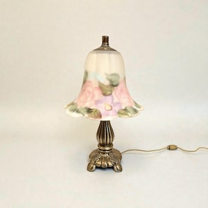 May include: A small, decorative table lamp with a floral design. The lamp features a cream-colored, bell-shaped shade adorned with pink and purple flowers and green leaves. The base is a bronze-colored metal with a detailed, ornate design. The lamp has a cord and plug.
