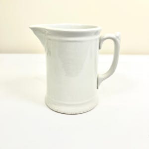 May include: A white ceramic pitcher with a handle and a spout. The pitcher has a cylindrical body with a slightly flared base and a simple, classic design. It is suitable for serving liquids.