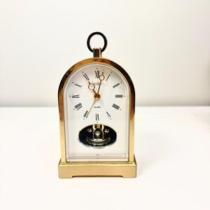 May include: A gold-toned mantel clock with an arched top and a circular handle. The white clock face features black Roman numerals, gold hands, and the text Linden and Quartz. A visible pendulum swings within a lower oval cutout window.