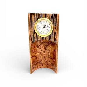 May include: A wooden desk clock with a gold-colored clock face and Roman numerals. The clock is set in a wooden frame with a curved base. The wood has a natural grain pattern with dark and light brown tones.