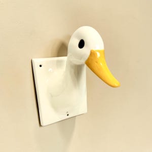 May include: A white ceramic wall hook shaped like a duck head. The duck has a smooth white finish, a bright yellow beak, and a single black eye visible on the side. The head is attached to a square white mounting plate with two small screw holes.