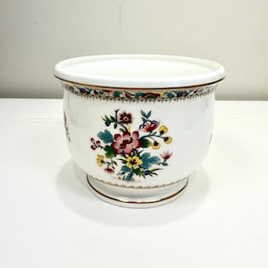 May include: A white ceramic planter with a floral design. The planter features a gold rim and base, and a colorful floral pattern with red, pink, yellow, and blue flowers. The planter is round and has a slightly flared shape.