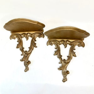 May include: Two ornate, gold-colored wall shelves with a scalloped top and intricate, scrolling leaf design. The shelves are made of a sturdy material and are designed to hold small decorative items. The shelves are a vintage style.