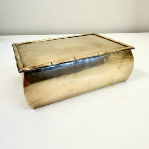 May include: A rectangular brass box with a smooth, curved body and a flat lid. The lid features a decorative border designed to resemble bamboo stalks. The metal surface has a polished, reflective finish with subtle signs of natural aging.