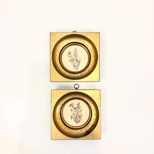 May include: Two square wall frames with a distressed gold finish and circular inner borders. Each frame contains a circular print featuring a detailed black and white botanical illustration of flowers, centered within a cream-colored background.