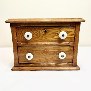 May include: A small, antique wooden chest of drawers with two drawers. The chest is made of light brown wood and features white, round knobs. A keyhole is visible on the top drawer. The chest has a simple, classic design.