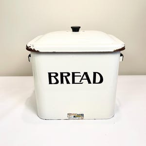 May include: A white bread box with a lid and a black knob. The word "BREAD" is printed in large black letters on the front. The box has a rectangular shape and appears to be made of metal. The edges show signs of wear.