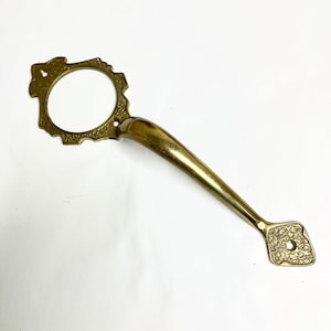 May include: A vintage brass door handle with an ornate design. The handle features a circular grip with a decorative edge, a curved handle, and a diamond-shaped end with a floral pattern. The brass has a warm, golden hue.