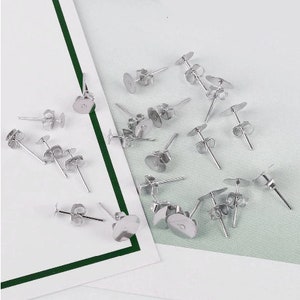 Może przedstawiać: Zbliżenie na grupę srebrnych sztyftów i zapięć do kolczyków. Sztyfty do kolczyków mają około 2,5 cm długości i mają małą pętelkę na górze. Zapięcia do kolczyków są okrągłe i mają mały otwór w środku.