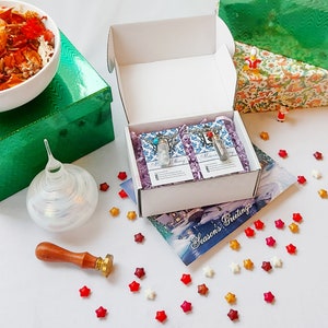 May include: A white box containing two small glass vial necklaces with blue and red accents, resting on a card that says "Season's Greetings". The box is surrounded by star-shaped candies and other decorative items.