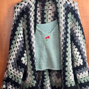 May include: A handmade crochet cardigan in a granny square pattern, featuring black, gray, and green squares. A light green knitted top with a small red flower embroidery is visible underneath the cardigan. The clothing is hanging on a metal hanger.